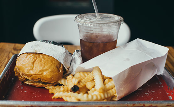 A meal containing paper bag filled with deep fried crinckle cut chips, burger and cola drink in plastic takeaway cup with lid and straw.